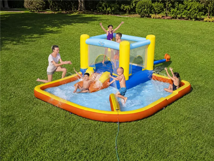 A large inflatable backyard splash pad and bouncy castle set up on a green lawn, with several children playing in the water and one standing in the bouncy top. The product is yellow, blue, and orange, and a hose is connected to the pool.